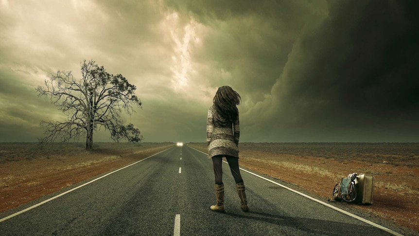 Lady standing on highway, storm in sky, tree on left, bags on right, oncoming car