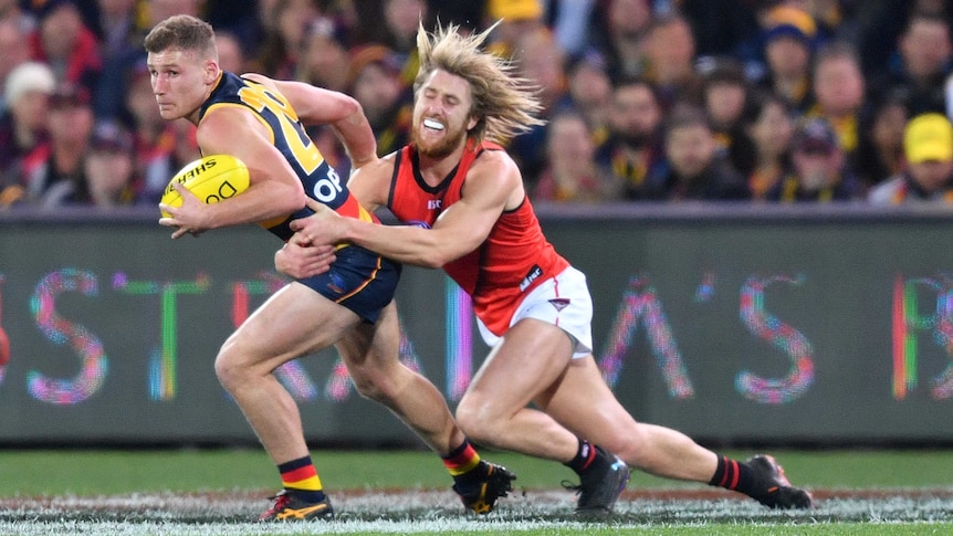 A male AFL player haolds the ball with his left hand as he is tackled around the waist by an opposition player.