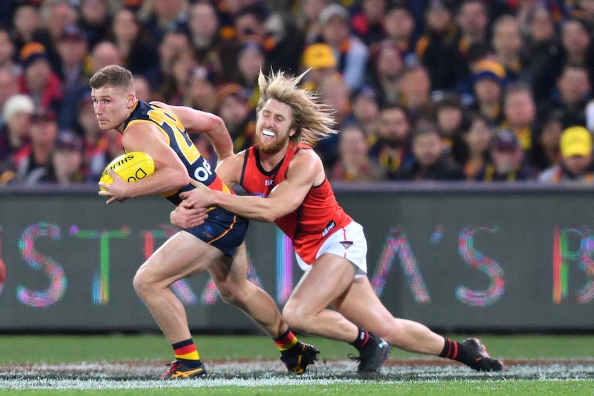 A male AFL player haolds the ball with his left hand as he is tackled around the waist by an opposition player.