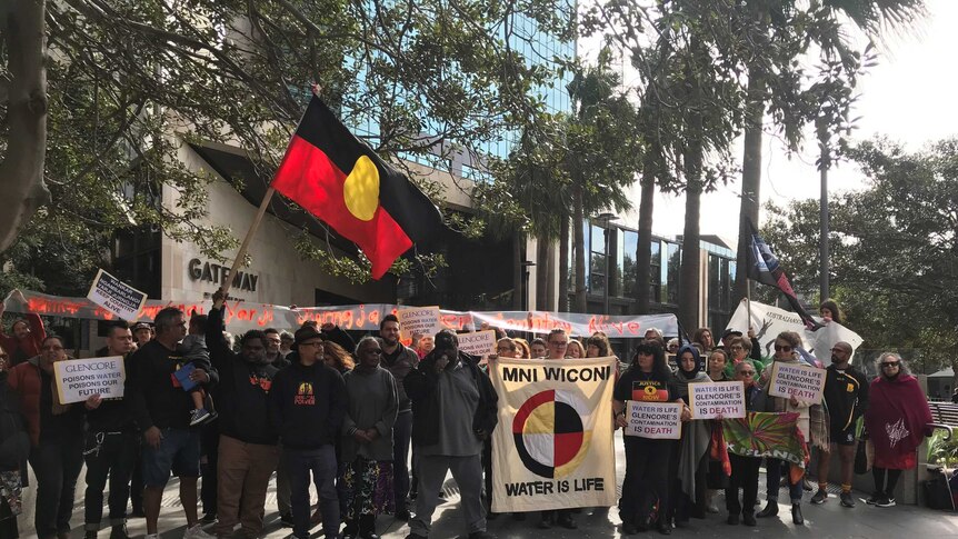About 20 people stand together, holding an Indigenous flag and signs.