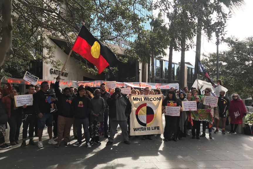 About 20 people stand together, holding an Indigenous flag and signs.