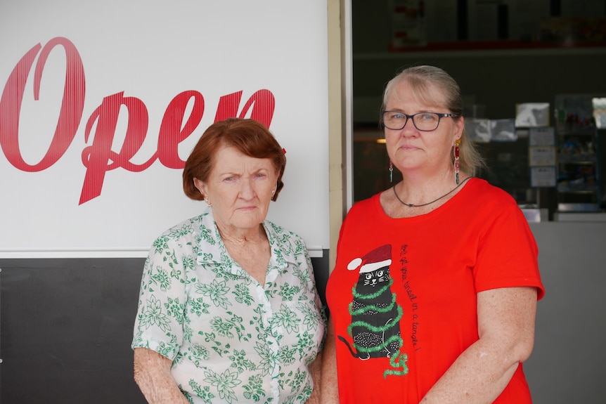 An older woman and a younger, taller, fair-haired woman stand outside a shopfront, looking angry.