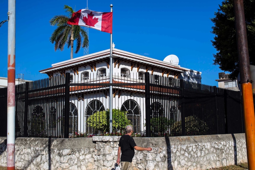 A man walks in front of a colonioal style white building, which is surrounded by the fence and has a canadian flag out the front