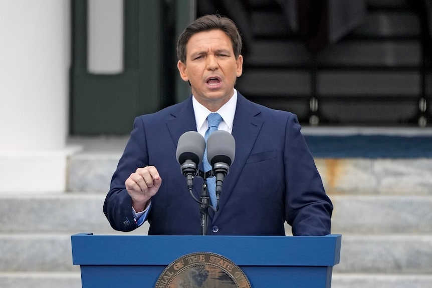 A middle-aged man with brown hair in a blue suit gestures emphatically while speaking behind a lectern outside.