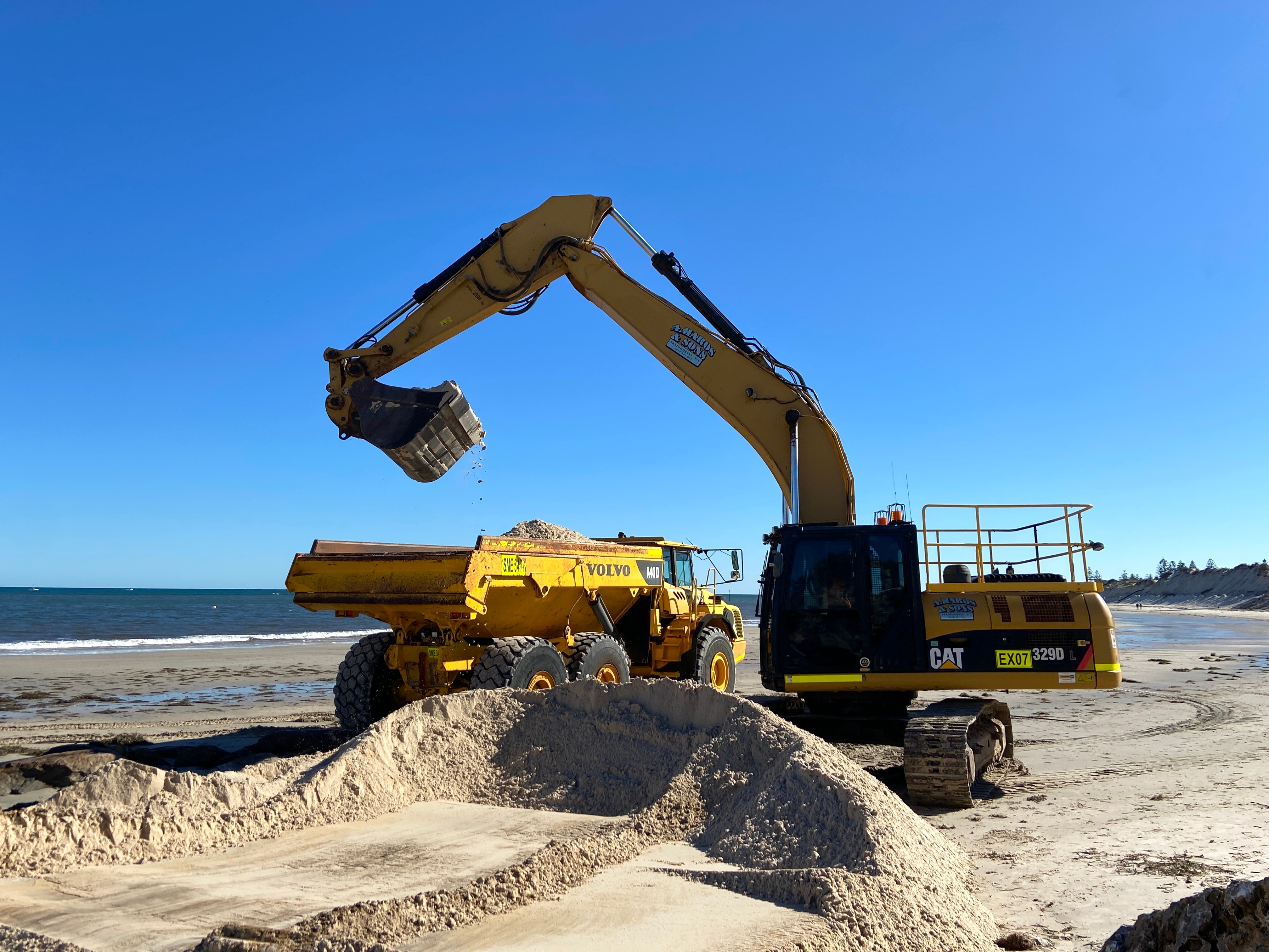 An excavator removes sand from a truck on a beach