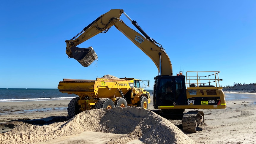 An excavator removes sand from a truck on a beach