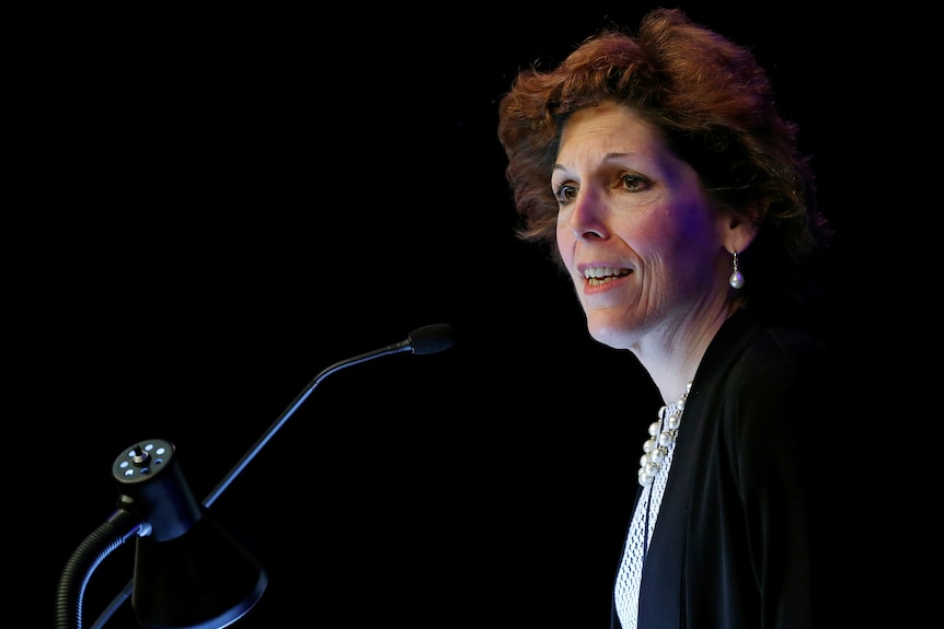 A woman with short brown hair wearing a black suit speaking into a microphone against a black background