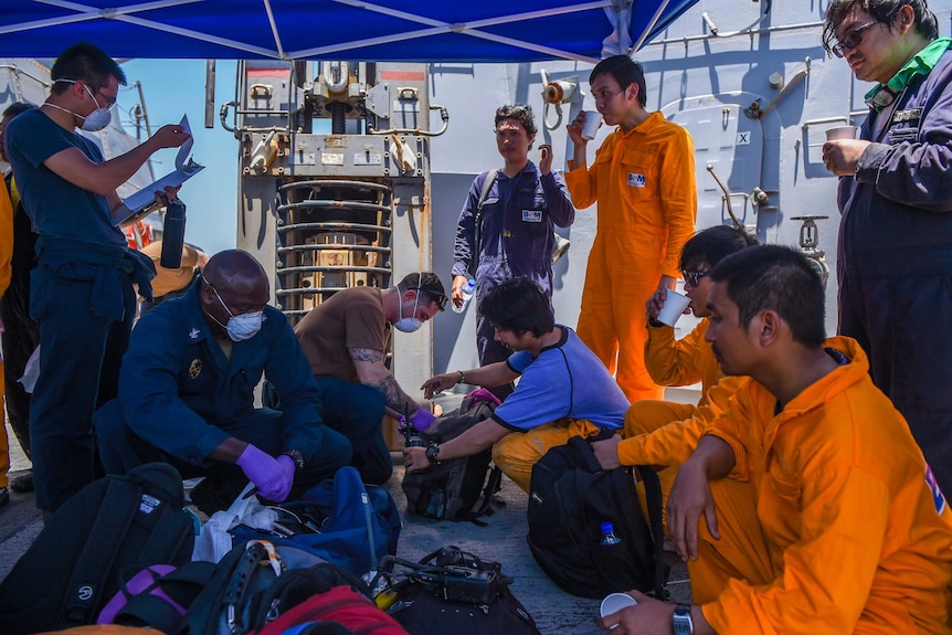 American sailors wearing face masks and latex gloves provide assistance to sailors wearing orange jumpsuits aboard a ship