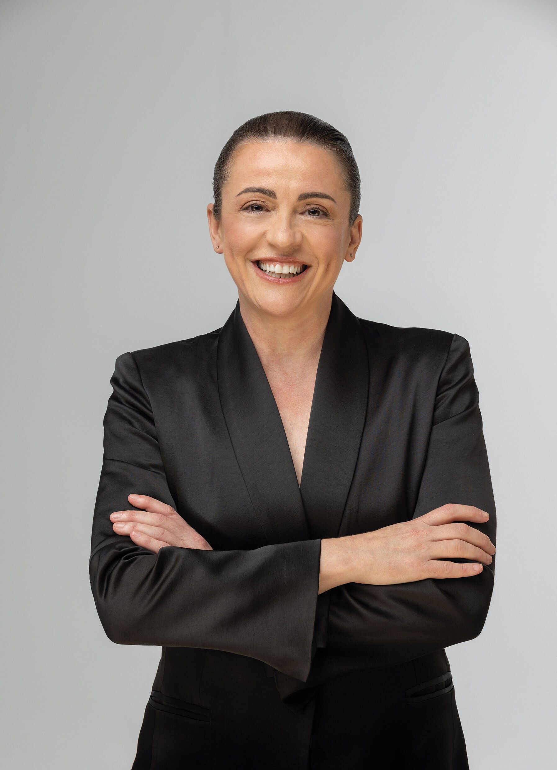 A middle-aged brown-haired woman smiles brightly. Her hair is pulled back severely, and her arms are folded.