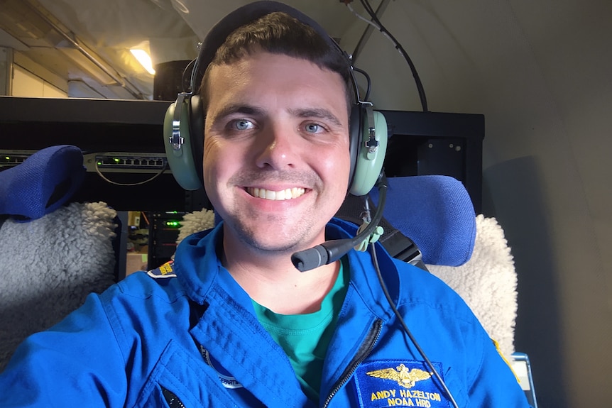 A man with big blue eyes smiles broadly while strapped into an aircraft seat, while wearing headphones.