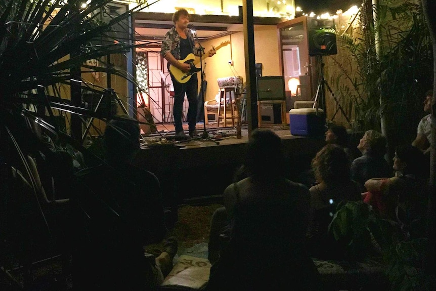 Dan Kelly playing guitar and singing on a back porch while people look on from the backyard