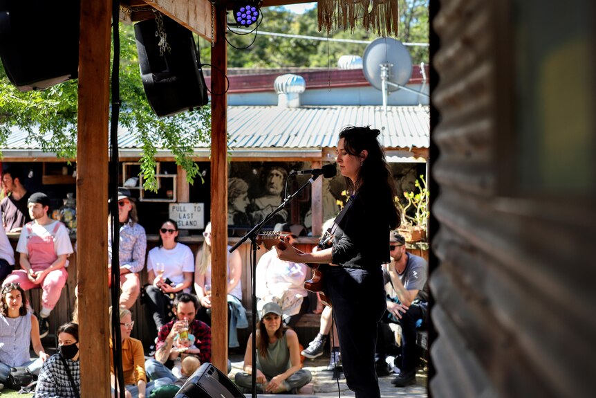 Woman with black hair on small stage holding guitar with seated outdoor audience watching on 