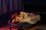 Two men in white robes entwined and kissing on a couch on stage