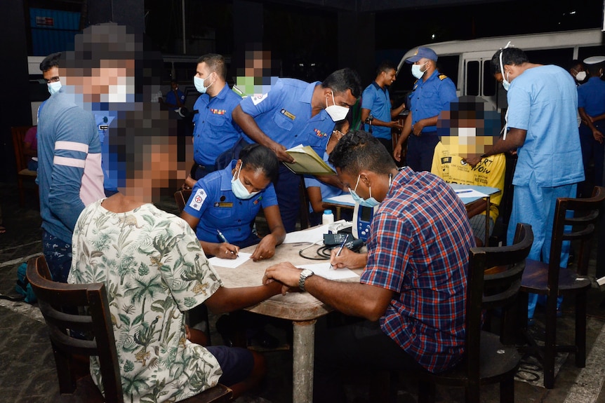 A group of people sit at a table surrounded by officials dressed in blue.