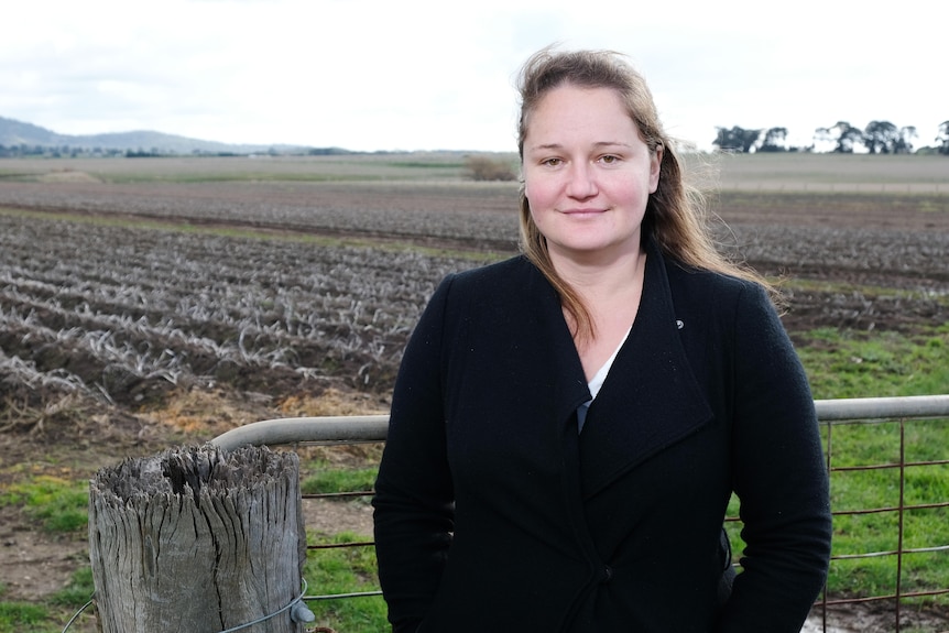 A woman with long, dark hair, wearing a dark jumper, stands in front of a potato field.