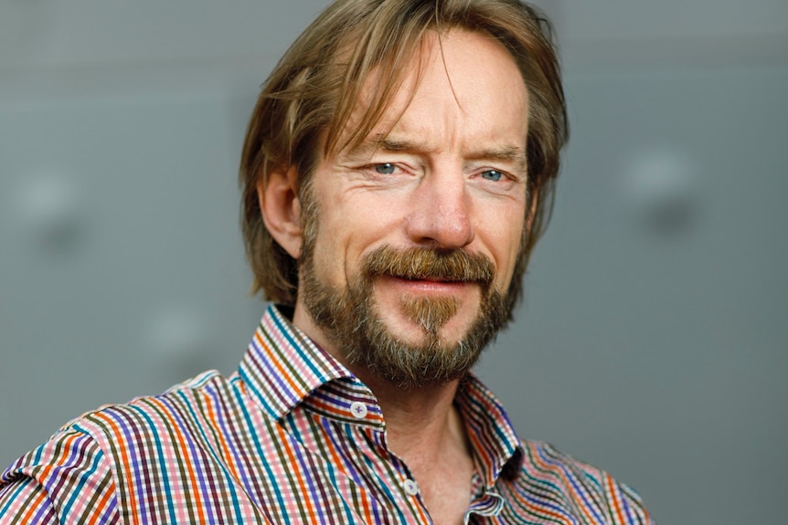 Jono Lineen, with fair hair and facial hair, in check shirt, sits with arms crossed smiling.