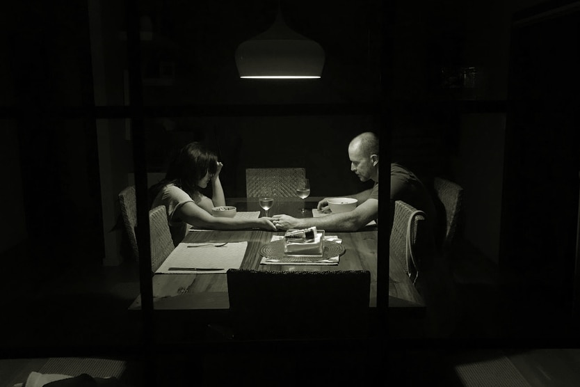 A black-and-white photo of a sad-looking man and woman holding hands across a dinner table.