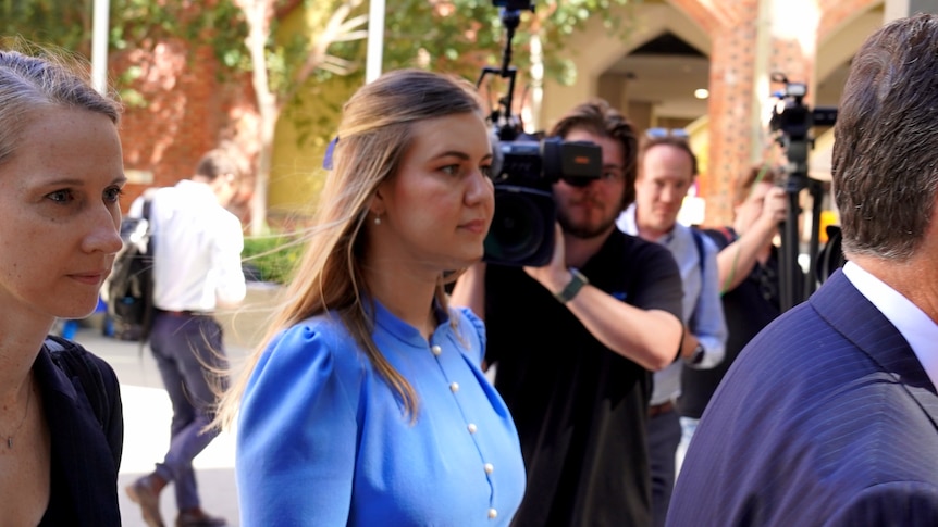A woman in a flowing blue dress arrives at a court building surrounded by cameras