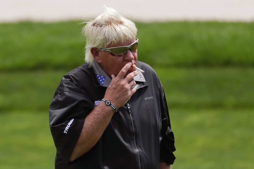 A man wearing sunglasses smokes a cigarette on a golf course.