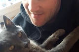 Close up photo of a young man in a beanie holding a grey cat with yellow eyes