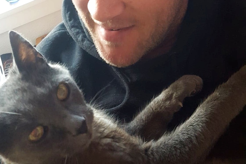 Close up photo of a young man in a beanie holding a grey cat with yellow eyes