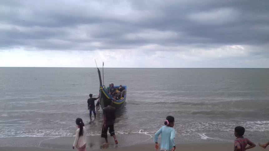 An Acehnese fishing boat lands as children stand on the beach under grey skies.