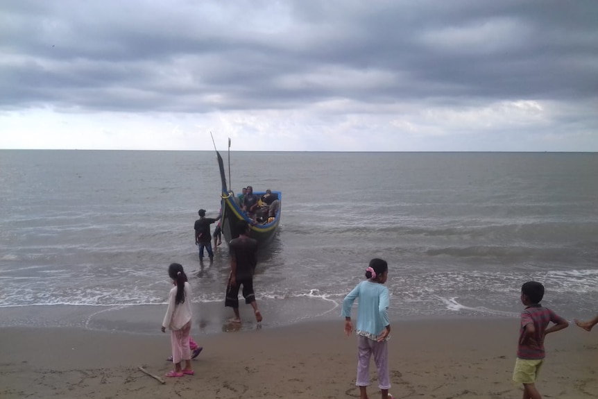 An Acehnese fishing boat lands as children stand on the beach under grey skies.