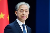 A Chinese man with silver hair speaks at a podium with a ed Chinese flag in the background with yellow stars.