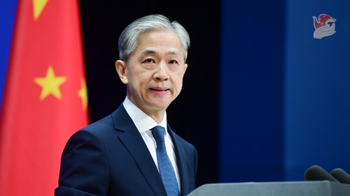 A Chinese man with silver hair speaks at a podium with a ed Chinese flag in the background with yellow stars.