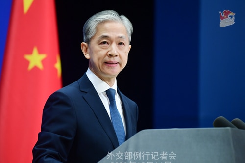 A Chinese man with silver hair speaks at a podium with a ed Chinese flag in the background with yellow stars.
