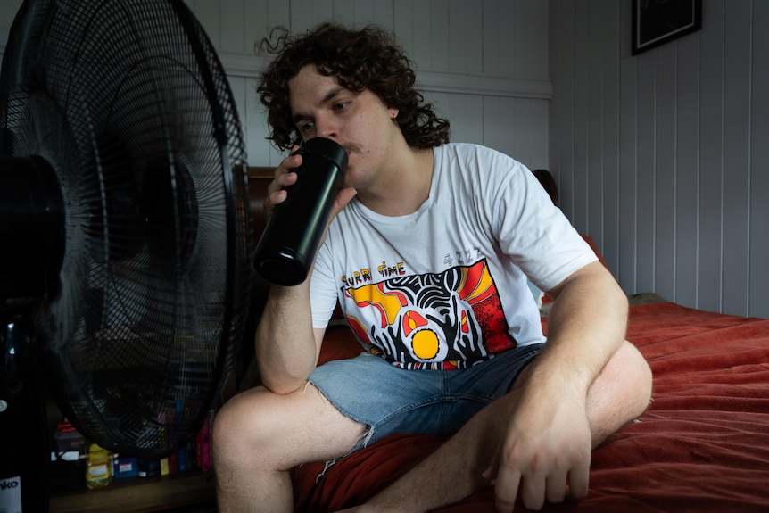 A young man sits in front of a fan on his bed drinking from a water bottle. 