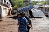 Person holds woman who is emotional behind them is debris of homes 