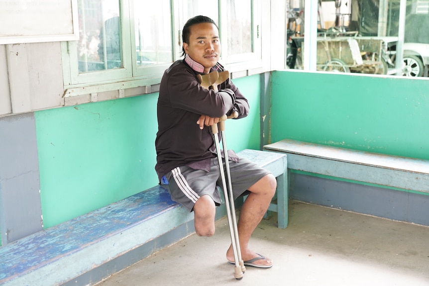 一个腿被截肢的男子坐在绿色长凳上，双臂拄着拐杖