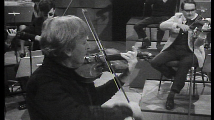 Yehudi Menuhin in black turtle-neck top plays his violin with members of the Menuhin Festival Orchestra playing on risers behind