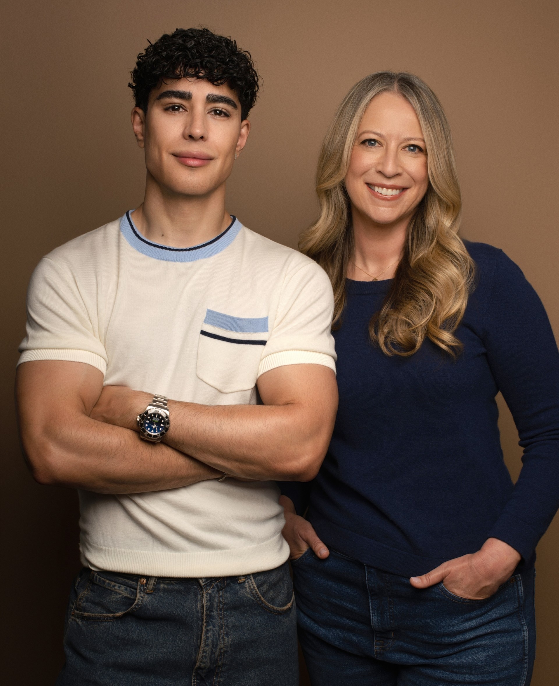 Un joven de cabello oscuro y rizado con una camiseta blanca y una mujer de mediana edad con cabello largo y rubio con una blusa de manga larga.