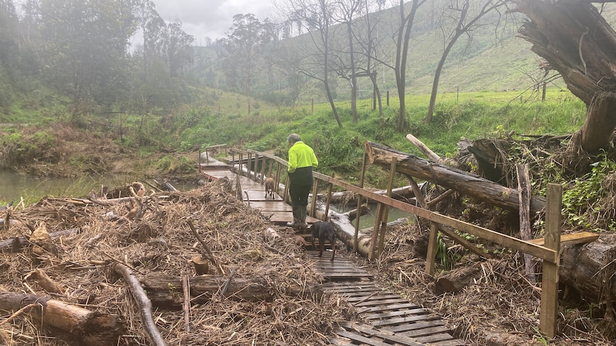 A man in high-vis crosses a makeshift bridge on a sodden country property.
