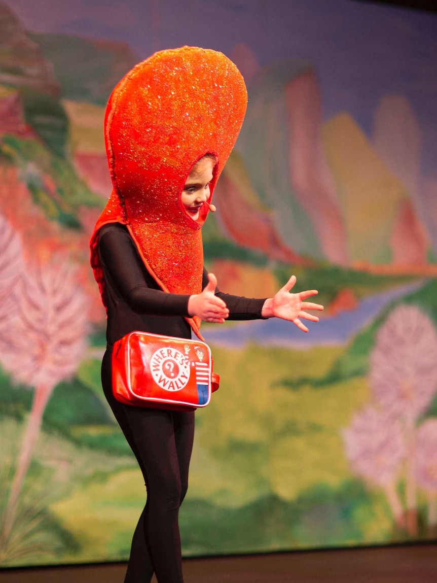 a girl on stage with a bright orange head-dress and handbag with arms outstretched to an audience