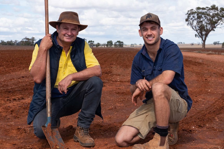 Two men crouch side by side in a paddock with one holding a shovel