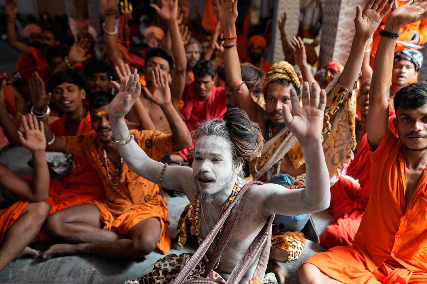 Hindu holy men in orange robes hold their hands up as they shout religious slogans while seated in a large group.