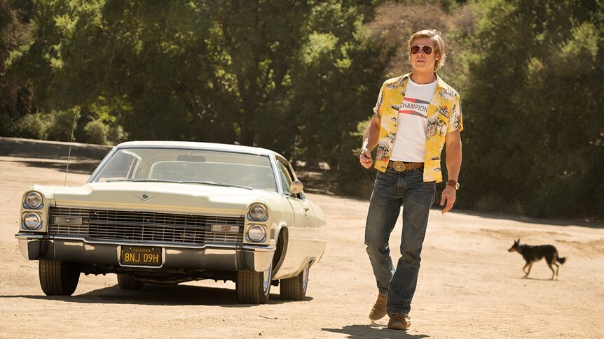 A man wearing bright yellow shirt and sunglasses walks along dirt road with trees away from cream coloured vintage car and dog.