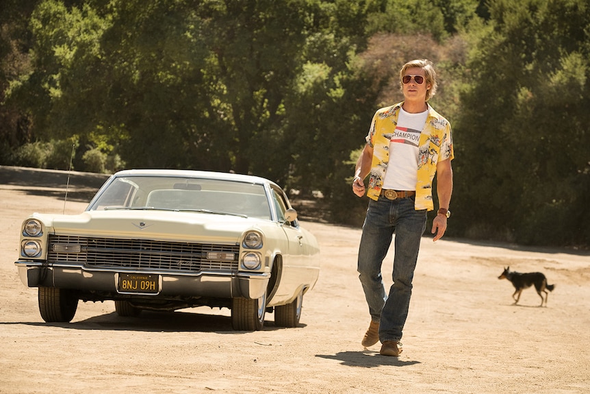 A man wearing bright yellow shirt and sunglasses walks along dirt road with trees away from cream coloured vintage car and dog.