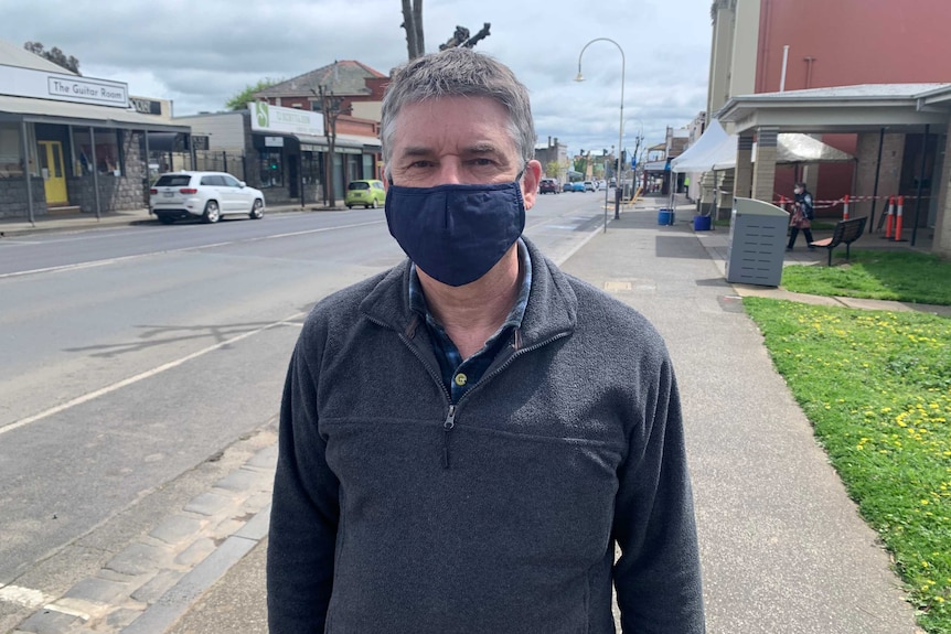 A man wearing a mask stands with Kilmore's main street behind him.