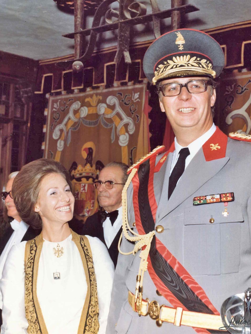 A tall man in military uniform smiles next to a young woman in white dress with stole around her neck