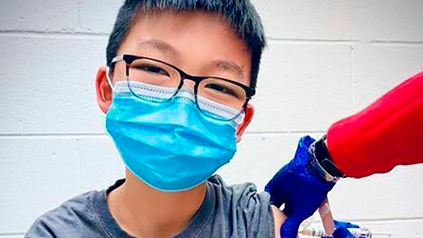 A young boy wearing glasses and a mask looks at the camera as he is vaccinated