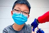 A young boy wearing glasses and a mask looks at the camera as he is vaccinated