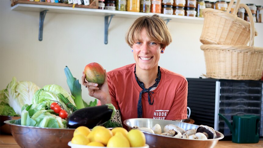 Hidden Harvest founder Berbel Franse holds a mango in front of bowls of fresh fruit and vegetables.