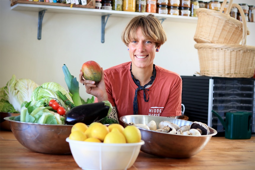 Hidden Harvest founder Berbel Franse holds a mango in front of bowls of fresh fruit and vegetables.