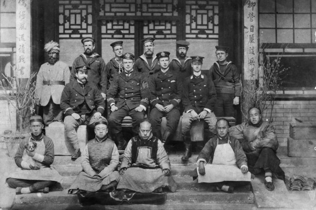 Black and white image of group of white men standing wearing military uniforms, and group of Chinese men seated before them.