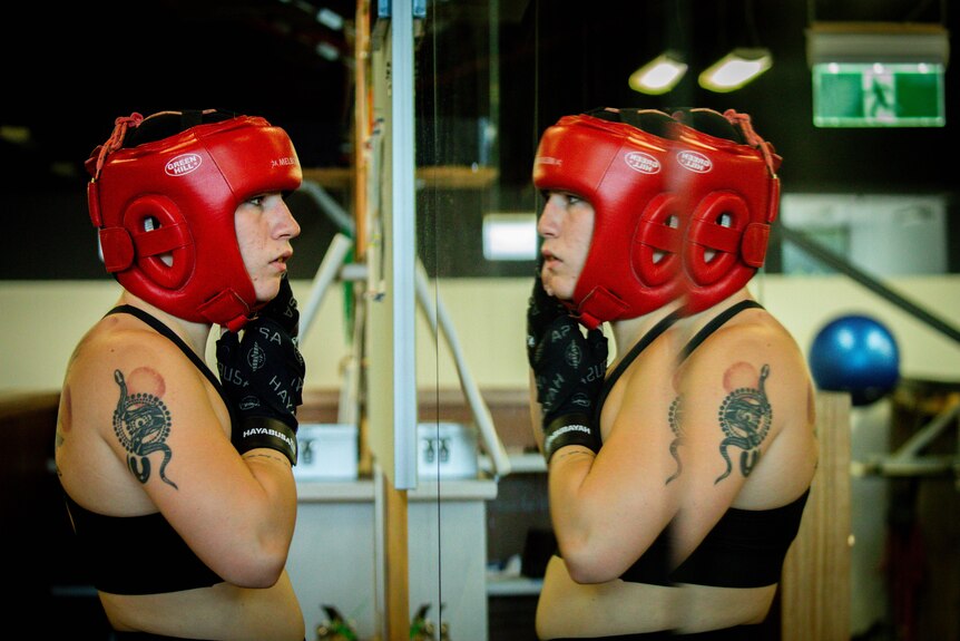 Boxer Marissa Williamson-Pohlman, tightening a red head protector to their chin, looking in a mirror at the gym