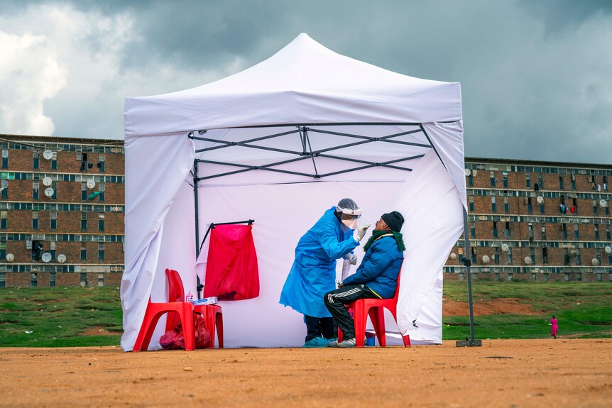 A health worker in full PPE takes a throat swab from a man in a white tent, with a building behind them under a stormy sky
